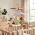 Handmade personalized felt baby mobile featuring dinosaurs, dinosaur eggs, and volcanoes, with the name 'LEO' hanging over a wooden baby crib, prehistoric-themed nursery decor