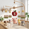 Handmade personalized felt baby mobile featuring a central red apple with the initial 'E', surrounded by farm animals including a dog, cow, pig, duck, and cat, hanging over a wooden baby crib, farmhouse-themed nursery decor.