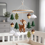 Handmade felt baby mobile featuring a central deer, surrounded by pine trees, oak leaves, acorns, and clouds hanging from a circular wooden frame over a baby crib, rustic forest nursery decor