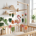 Handmade felt flamingo baby mobile with hibiscus flowers and tropical leaves hanging above a wooden crib.
