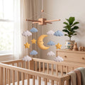 Handmade felt baby mobile featuring a sleepy crescent moon, stars, and fluffy clouds in pastel colors hanging over a wooden baby crib, dreamy sky-themed nursery decor