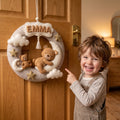 Personalized felt wreath with a sleeping teddy bear on a crescent moon, clouds, stars, and the name Emma, hanging on a wooden door.