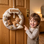Personalized felt wreath with a sleeping teddy bear on a crescent moon, clouds, stars, and the name Emma, hanging on a wooden door.