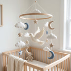 Handmade felt baby mobile featuring a white dove, crescent moons, stars, and fluffy clouds hanging from a circular wooden frame over a baby crib, peaceful nursery decor.