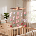 Handmade personalized felt baby mobile featuring white bunnies, pink tulips, butterflies, strawberries, and a rainbow, with the name 'EMMA' hanging over a wooden baby crib, nursery decor