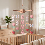 Handmade personalized felt baby mobile featuring white bunnies, pink tulips, butterflies, strawberries, and a rainbow, with the name 'EMMA' hanging over a wooden baby crib, nursery decor