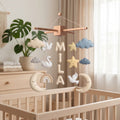Handmade personalized felt baby mobile with white swans, peace doves, rainbows, and crescent moons, featuring the name 'MILA' hanging over a wooden baby crib, dreamy nursery decor.